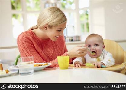 Mother Feeding Baby Sitting In High Chair At Mealtime