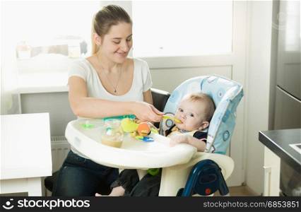 Mother feeding her 9 months old baby son sitting in child chair on kitchen