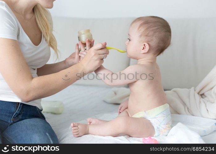 Mother feeding her adorable baby boy on bed with fruit sauce from glass jar