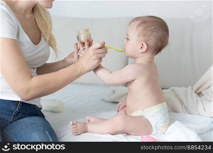 Mother feeding her adorable baby boy on bed with fruit sauce from glass jar