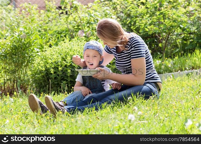Mother feeding her young son on the grass in the garden as they relax together in the sunshine