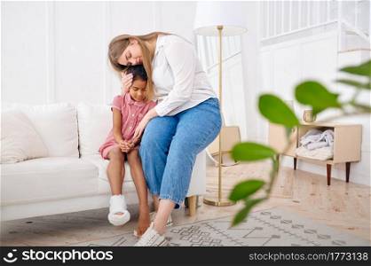 Mother feels sorry for her little daughter on sofa in living room.Mom and female child leisures in their house together, good relationship, parental care and love. Mother feels sorry for her little daughter