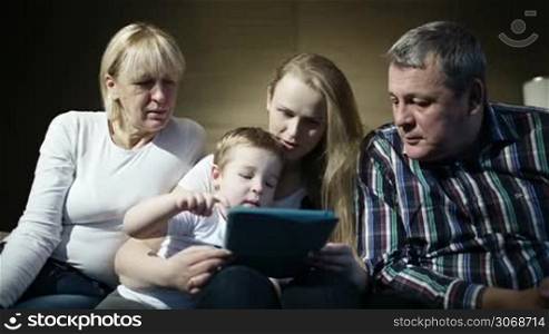 Mother, grandmother and granfather watching boy playing game on touchpad. Grandpa trying to help