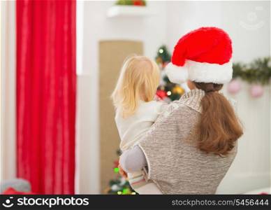 Mother holding baby and looking on Christmas tree. Rear view
