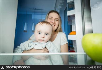 Mother holding baby and taking green apple from apple. View from inside of refrigerator
