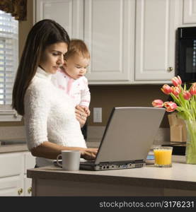 Mother holding baby and typing on laptop computer in kitchen.