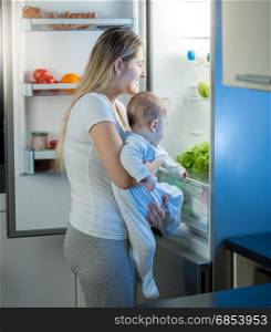 Mother holding baby son and looking inside of refrigerator at night