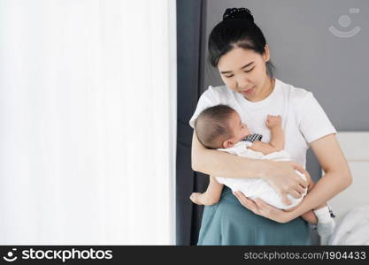 mother holding newborn baby sleeping in her arm near window in the bedroom