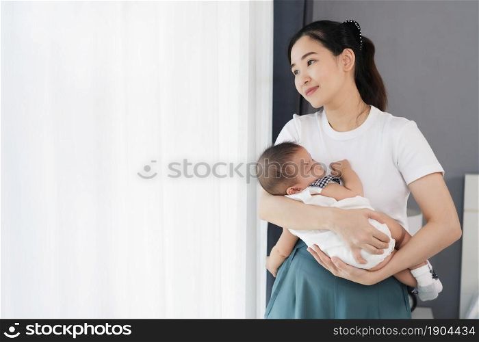 mother holding newborn baby sleeping in her arm near window in the bedroom