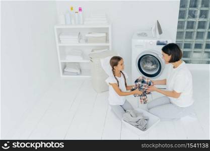 Mother housewife and her daughter load washing machine with dirty clothes, pose in spacious laundry room, does chores at home, look happily at each other. Laundry day concept. Family in washing room