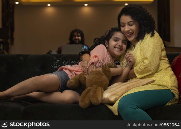 Mother hugging her daughter on sofa and father watching them from a distance