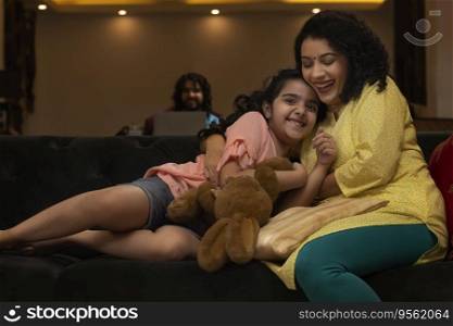 Mother hugging her daughter on sofa and father watching them from a distance