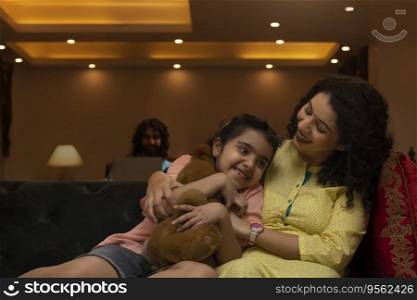 Mother hugging her daughter on sofa and father watching them from a distance
