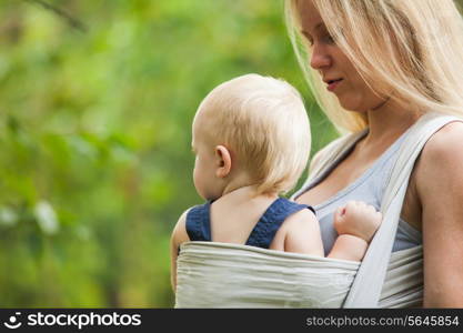 Mother is carrying her child and walking. Baby in sling outdoor.