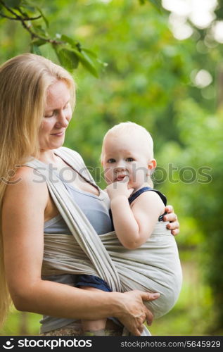 Mother is carrying her child and walking. Baby in sling outdoor.