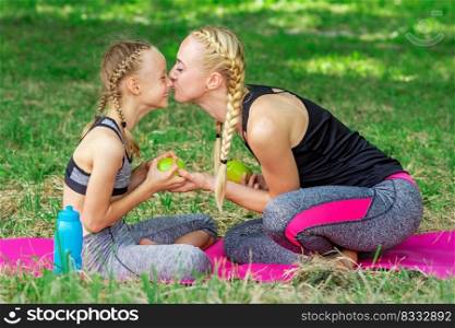 Mother kissing her daughter in nose giving green apple sitting on the grass in the park. Mother kissing her daughter in the park