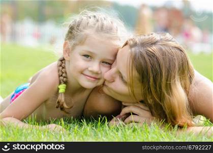 Mother kissing her daughter lying on a green grass lawn