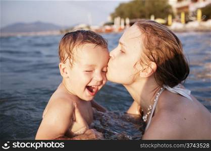 Mother kissing her young child while bathing in the sea waters during sunset