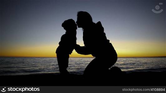 Mother kneels kissing child at waters edge, sunset