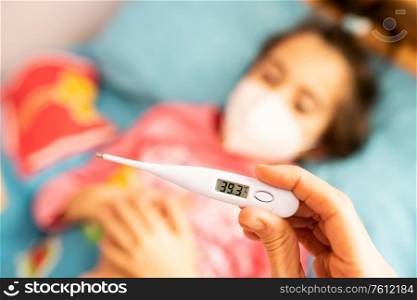 Mother measuring her daughter&rsquo;s temperature, protected by a mask, with a digital thermometer to check for fever. Mother measuring her toddler&rsquo;s temperature, protected by a mask.