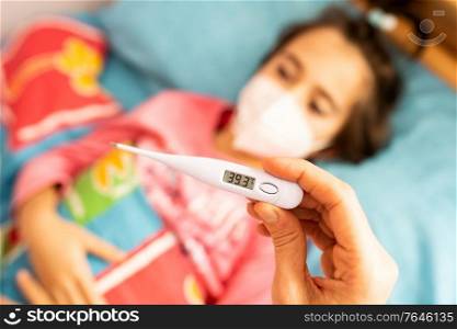 Mother measuring her daughter&rsquo;s temperature, protected by a mask, with a digital thermometer to check for fever. Mother measuring her daughter&rsquo;s temperature, protected by a mask.
