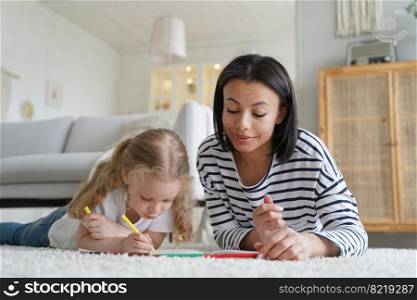 Mother or babysitter and adopted daughter draw together lying on floor carpet at home, mom with preschooler kid girl painting by colorful pencils. Child creativity development, adoption concept.. Mother and adopted daughter paint by colorful pencils, lying on floor at home. Creativity, adoption