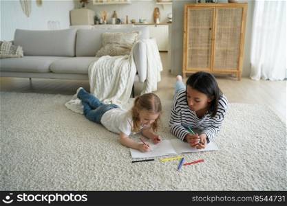 Mother or babysitter and little kid girl lying on floor carpet in living room drawing together, mom with daughter painting enjoy creative hobby. Child creativity development, parenting concept.. Mother, little daughter paint together lying on floor in living room. Child creativity, parenting