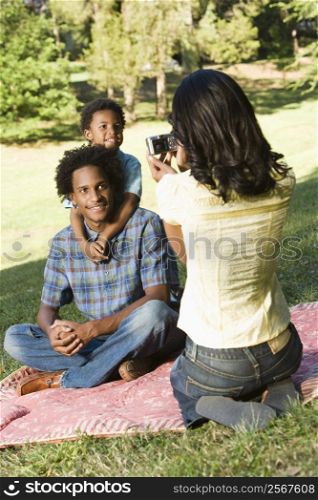Mother photographing son and husband with digital camera in park.