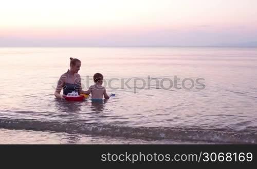 Mother playing with young son in the beach waters in the distance during sunset with red toy boat