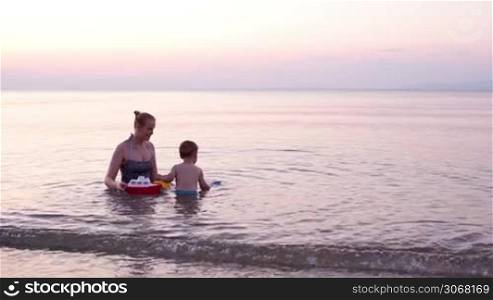 Mother playing with young son in the beach waters in the distance during sunset with red toy boat