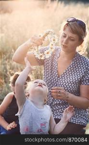 Mother putting a coronet of wild flowers on a head of little girl. Family spending time together on a meadow, close to nature. Parents and children playing together. Candid people, real moments, authentic situations
