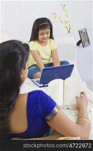 Mother reading a book while daughter plays with computer