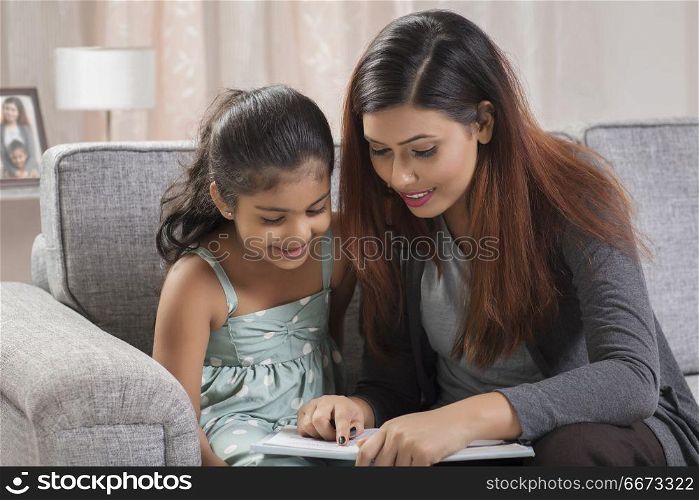 Mother reading book to daughter sitting on sofa at home
