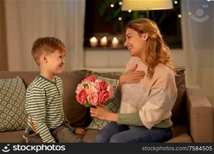 mother&rsquo;s day, holidays and family concept - happy little son giving flowers to his smiling mother at home in evening. smiling little son gives flowers to mother at home