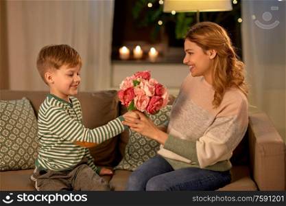 mother&rsquo;s day, holidays and family concept - happy little son giving flowers to his smiling mother at home in evening. smiling little son gives flowers to mother at home
