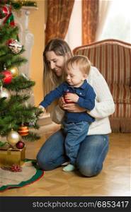 Mother showing her baby son how to decorate Christmas tree with baubles