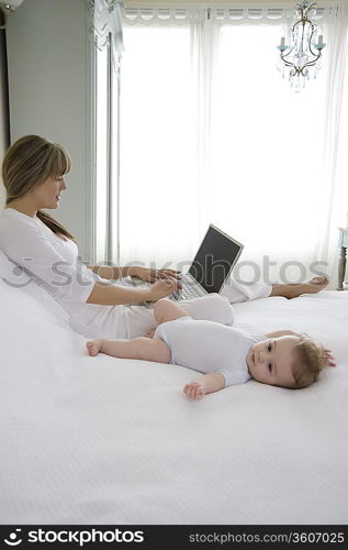 Mother sits typing on laptop with baby beside her on bed