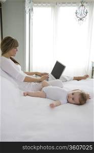 Mother sits typing on laptop with baby beside her on bed