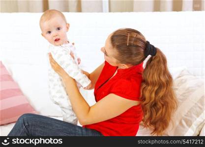 Mother sitting on divan and holding intersted baby in hands&#xA;
