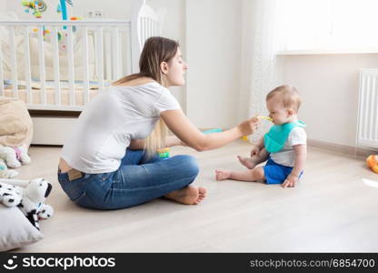 Mother sitting on floor at living room and feeding her baby with porridge