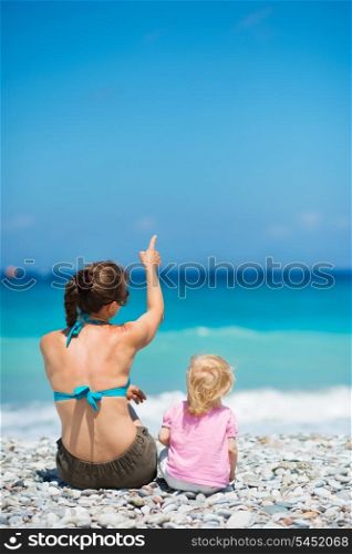 Mother sitting with baby on beach and pointing on copy space. Rear view