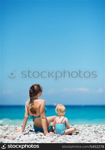 Mother sitting with baby on beach looking into distance. Rear view