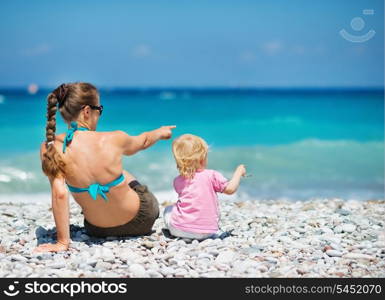 Mother sitting with baby on sea shore and pointing on copy space. Rear view