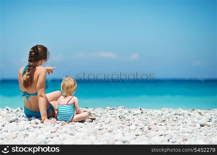 Mother sitting with baby on sea shore looking into distance. Rear view