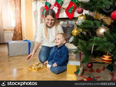 Mother sitting with her baby son on floor at living room next to fireplace and christmas tree