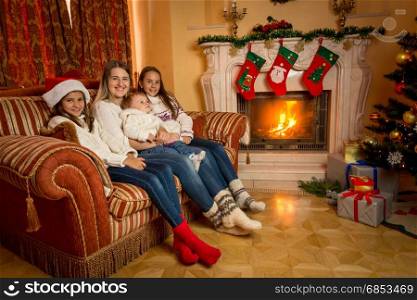 Mother sitting with her daughters on sofa living room next to burning fireplace on Christmas