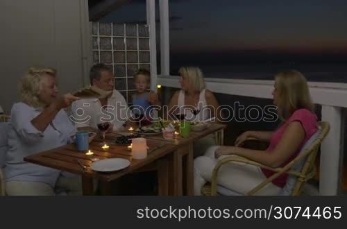 Mother, son and grandparents dining on the outdoor balcony late in the evening. Everyone taking bread and hen adults toasting with wine and drinking