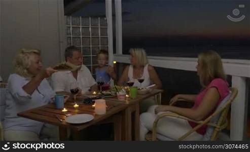 Mother, son and grandparents dining on the outdoor balcony late in the evening. Everyone taking bread and hen adults toasting with wine and drinking