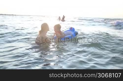 Mother swimming with her little boy in the sea standing chest high in the water holding his hands as he balances on a floating plastic ring