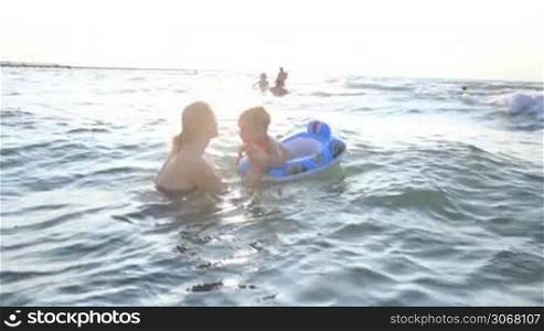 Mother swimming with her little boy in the sea standing chest high in the water holding his hands as he balances on a floating plastic ring